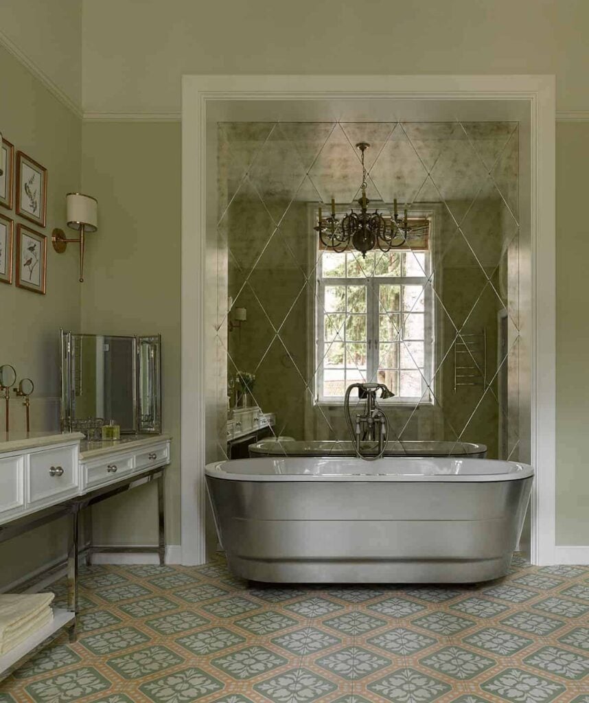 明亮的银色独立式浴缸是这间浴室的亮点，它有浅绿色图案的地砖，与梳妆台的米色台面相匹配，梳妆台的绿色墙壁装饰着框架艺术品和壁挂式灯具。