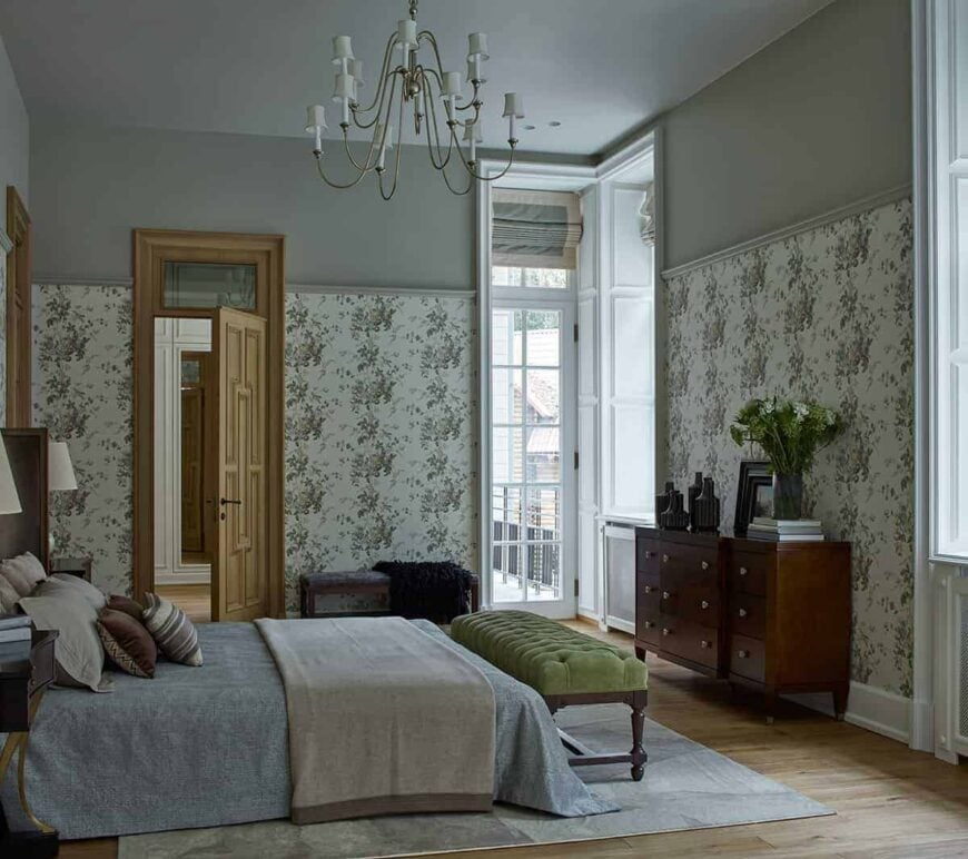 这间迷人的传统卧室有绿色的花墙，作为木制梳妆台的漂亮背景，与硬木地板相辅相成。灰色的床下面有一张浅灰色的地毯，床脚有一张绿色的软垫长凳。