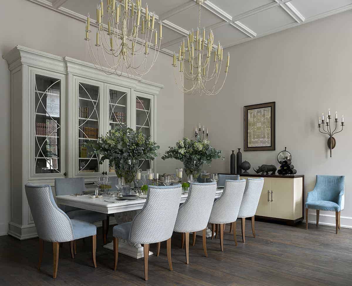 这是一个优雅而传统的正式餐厅，硬木地板与白色餐桌周围的软垫餐椅形成鲜明对比，白色餐桌与白色墙壁旁的白色橱柜相匹配。长桌上方有几盏白色细枝形吊灯，悬挂在白色格子天花板上。